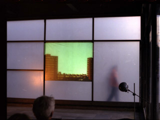 A still image of the performance In 24 Hours by Corali Dance Company, 2000. The photo shows the back end of an empty shop unit with frosted glass at one end of it and a film projected on the window of a city scape. Dancer John Long in an orange top walks past the window on the outside.